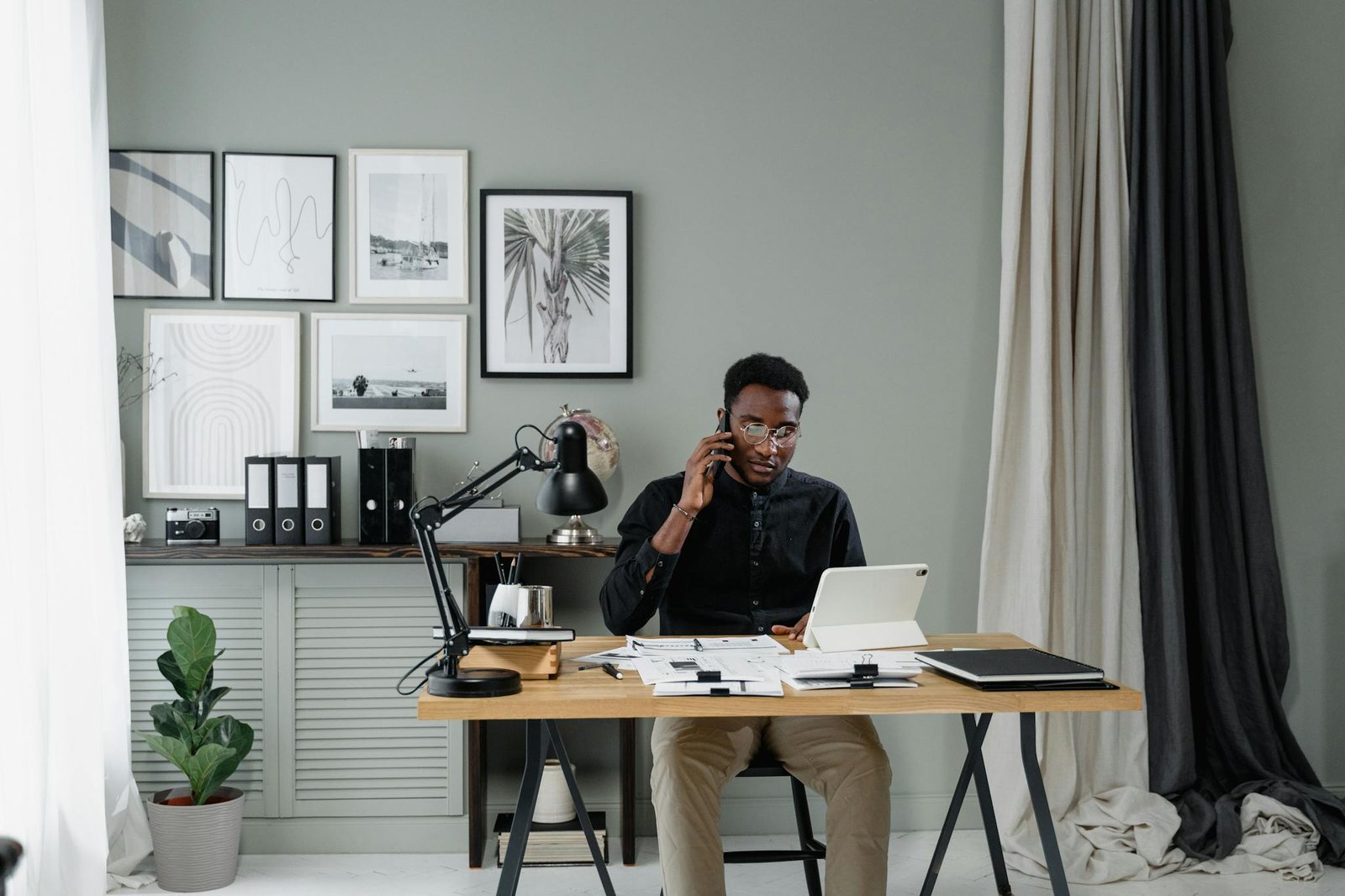 A Man in Black Long Sleeves Talking on the Phone while Sitting Beside the Wooden Table